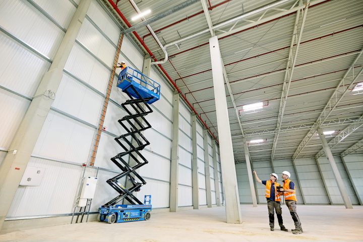 Arbeiter in Lagerhalle auf Hebebühne, diskutieren Bauarbeiten.